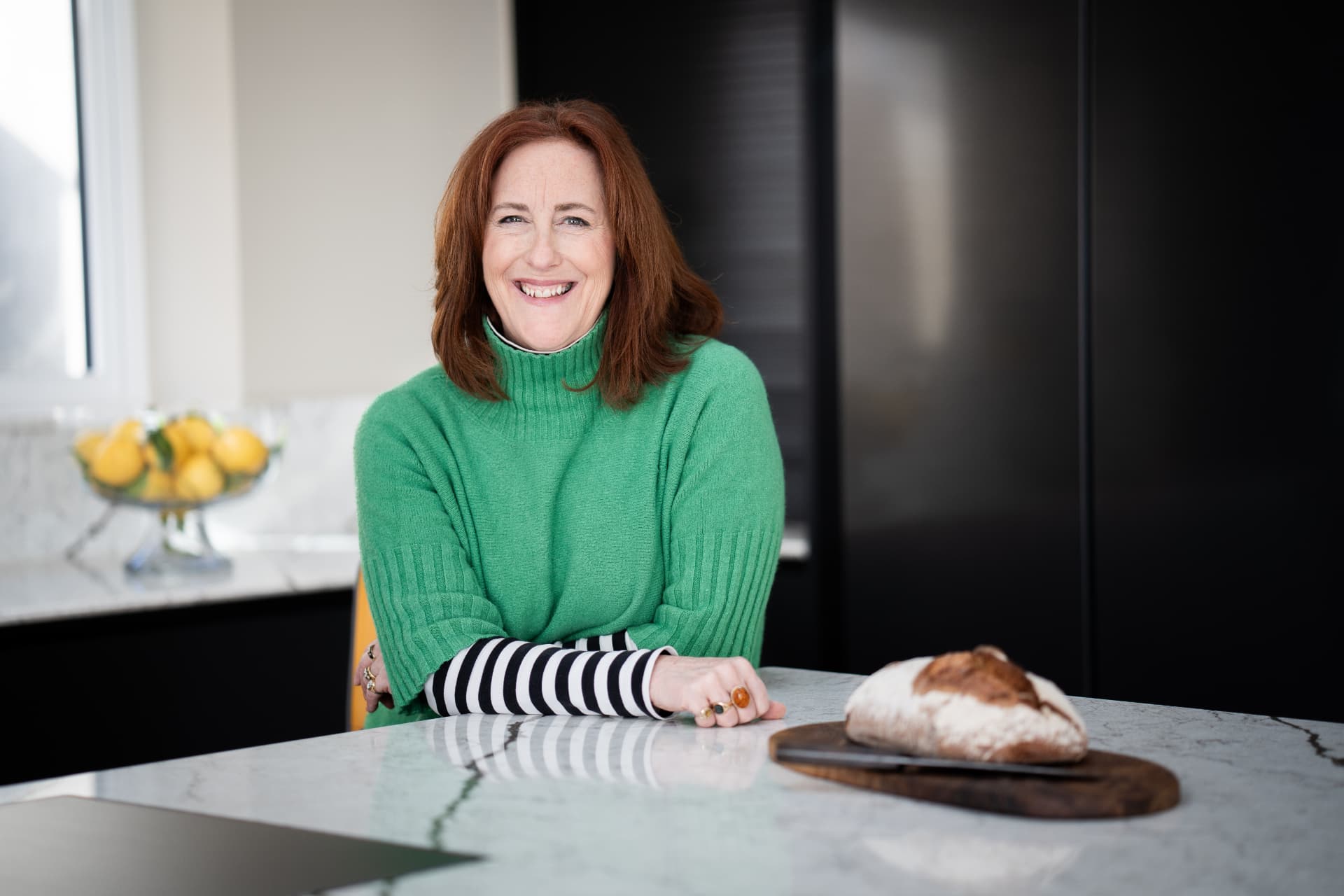 Tess Stobie at a kitchen island in a dark green shaker kitchen, smiling